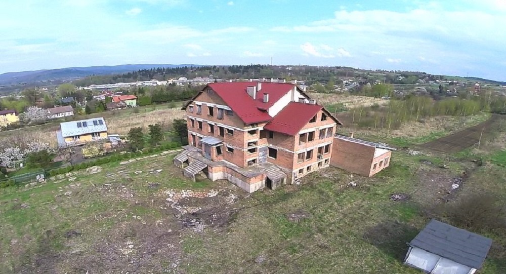 Niedokończony, ceglany budynek z czerwonym dachem na wzg&oacute;rzu. Widok z lotu ptaka, w tle zabudowa i zieleń.